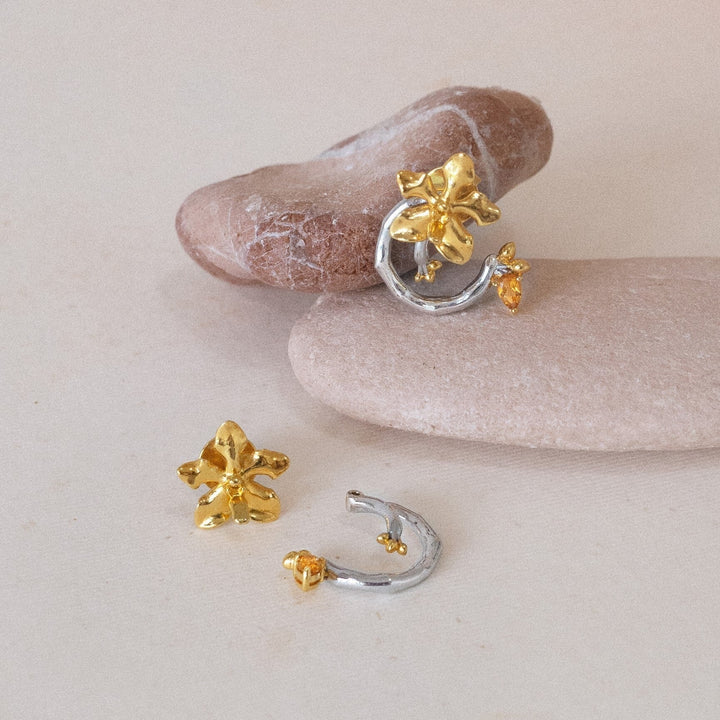 Risis Orchid Spiral Earrings - A slender hand adorned with elegant jewelry: a silver bangle bracelet featuring golden floral embellishments, paired with a matching ring, both showcasing intricate details and a warm, golden hue.