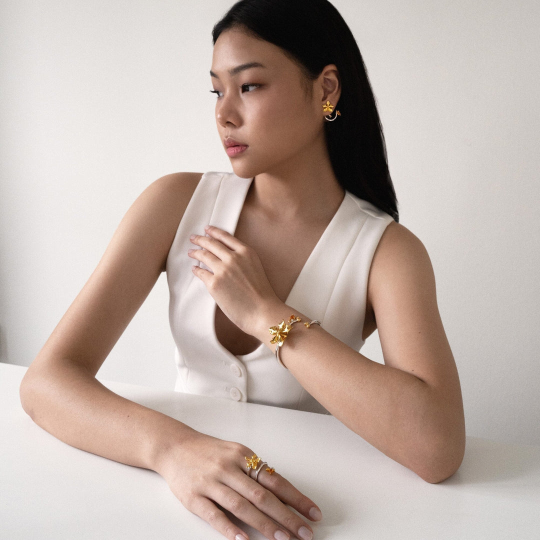 Risis Orchid Spiral Earrings - The image shows a gold-toned earring with intricate designs, featuring a small bee and flowers, complemented by warm, translucent gemstones, worn on an ear.