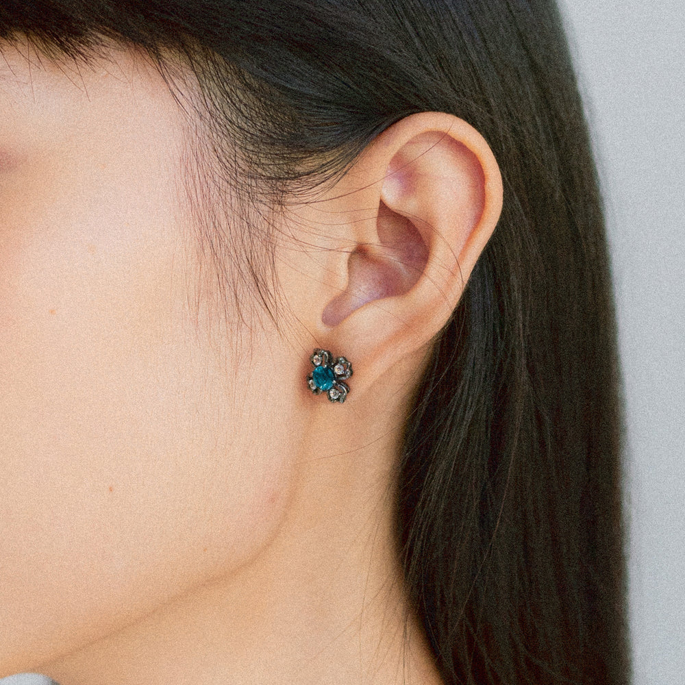 Risis Peranakan Kebaya Earrings (BRH) with London Blue Topaz - A dark blue, flower-shaped earring with sparkling petals adorns a person's earlobe, providing a touch of elegance.