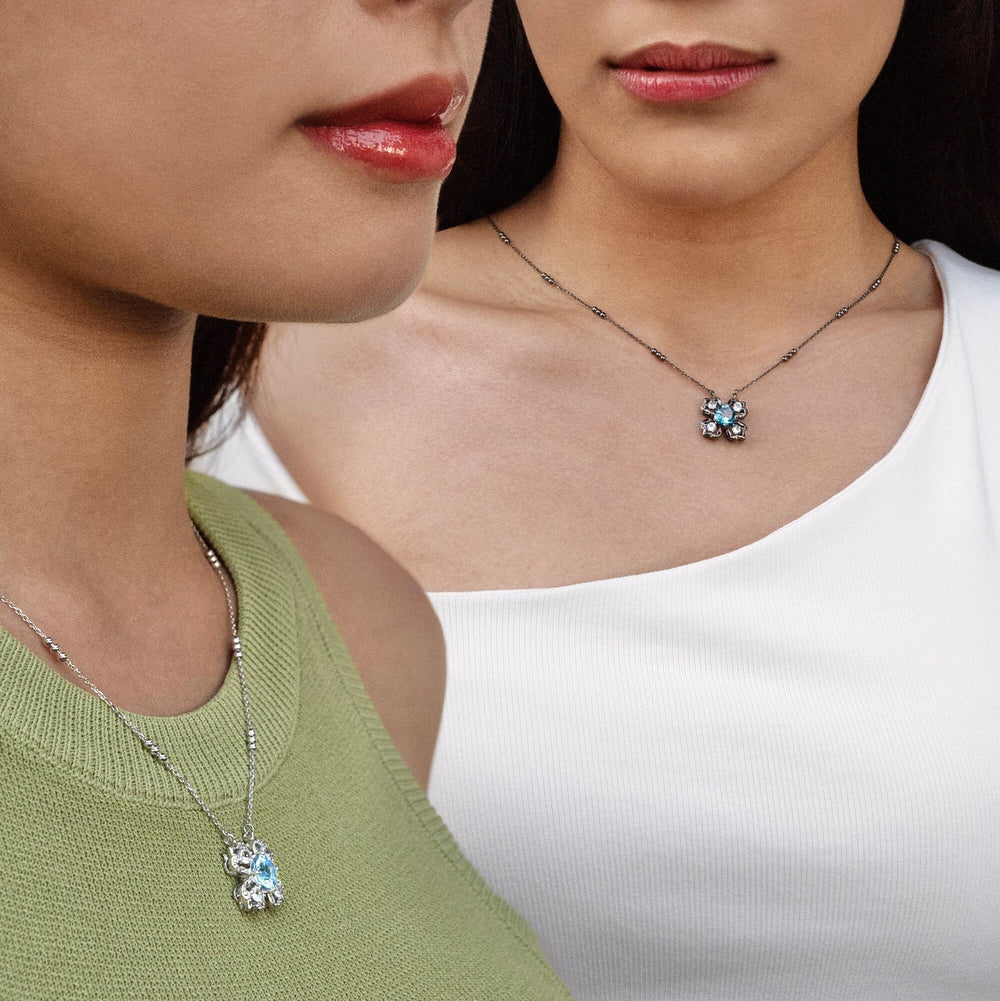 Risis Peranakan Kebaya Necklace (BRH) with London Blue Topaz - Two pendants on silver chains, one has a larger blue gem flanked by clear stones, the other a smaller blue stone with a sparkling halo, adorn two individuals' necklines.