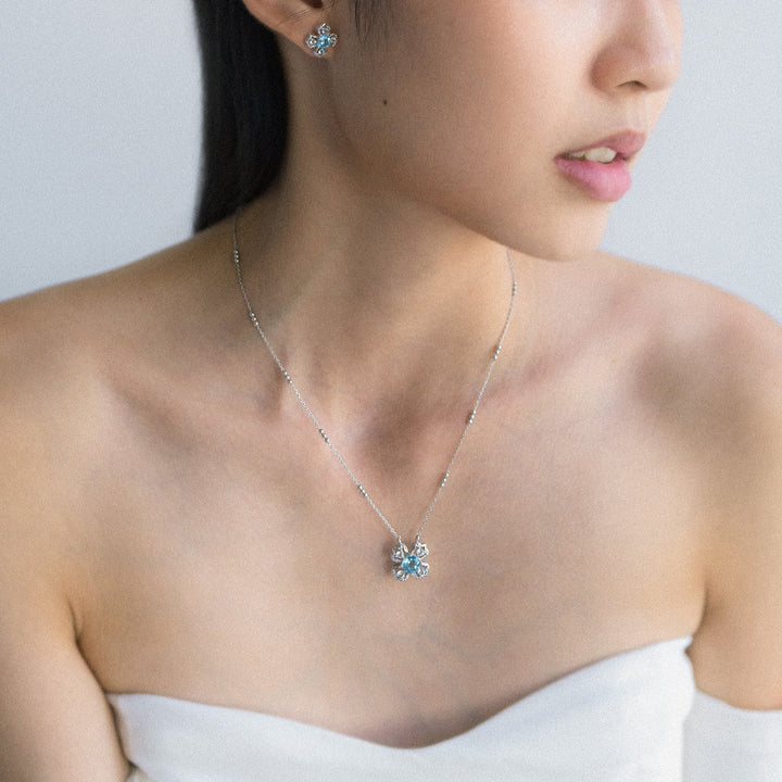 Risis Peranakan Kebaya Necklace (RH) with Sky Blue Topaz - A delicate silver necklace with a glittering blue gem pendant rests against a person's collarbone, complemented by matching earrings. The jewelry exudes elegance and simplicity.