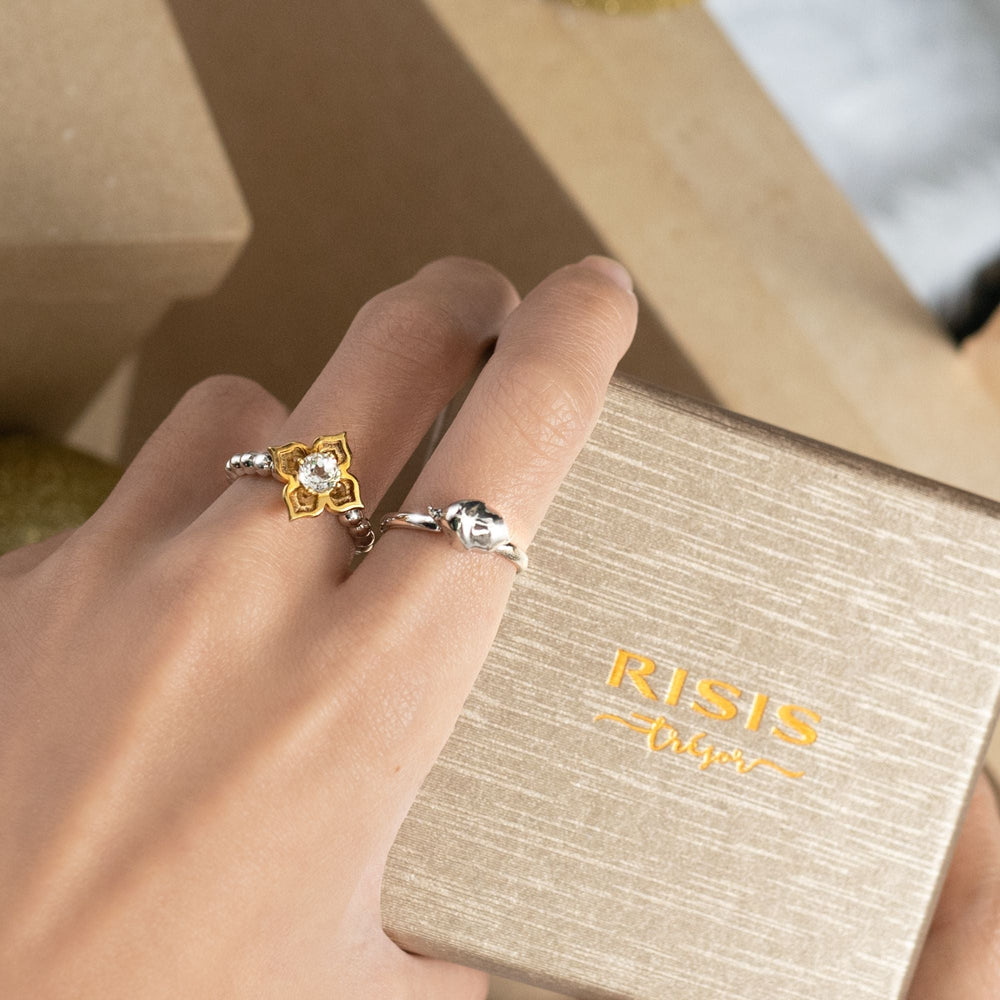 Risis Peranakan Moments Beadwork Ring with White Topaz - A hand displays two rings: one gold with a floral design and central gemstone, and another silver with a simpler band and single gemstone.