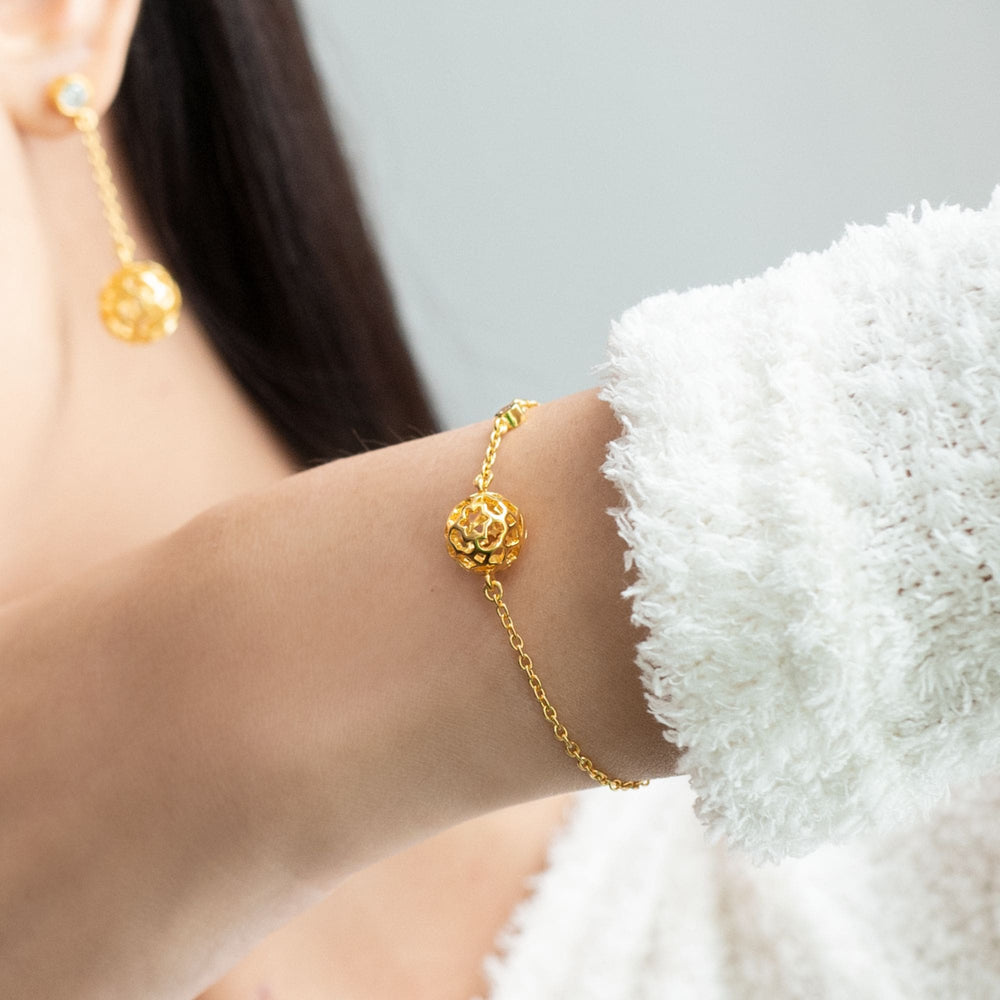 Risis Peranakan Spheres Bracelet with White Topaz (G) - A golden bracelet and matching earrings adorn a person's wrist and earlobe, featuring intricate spherical charms with a floral pattern.