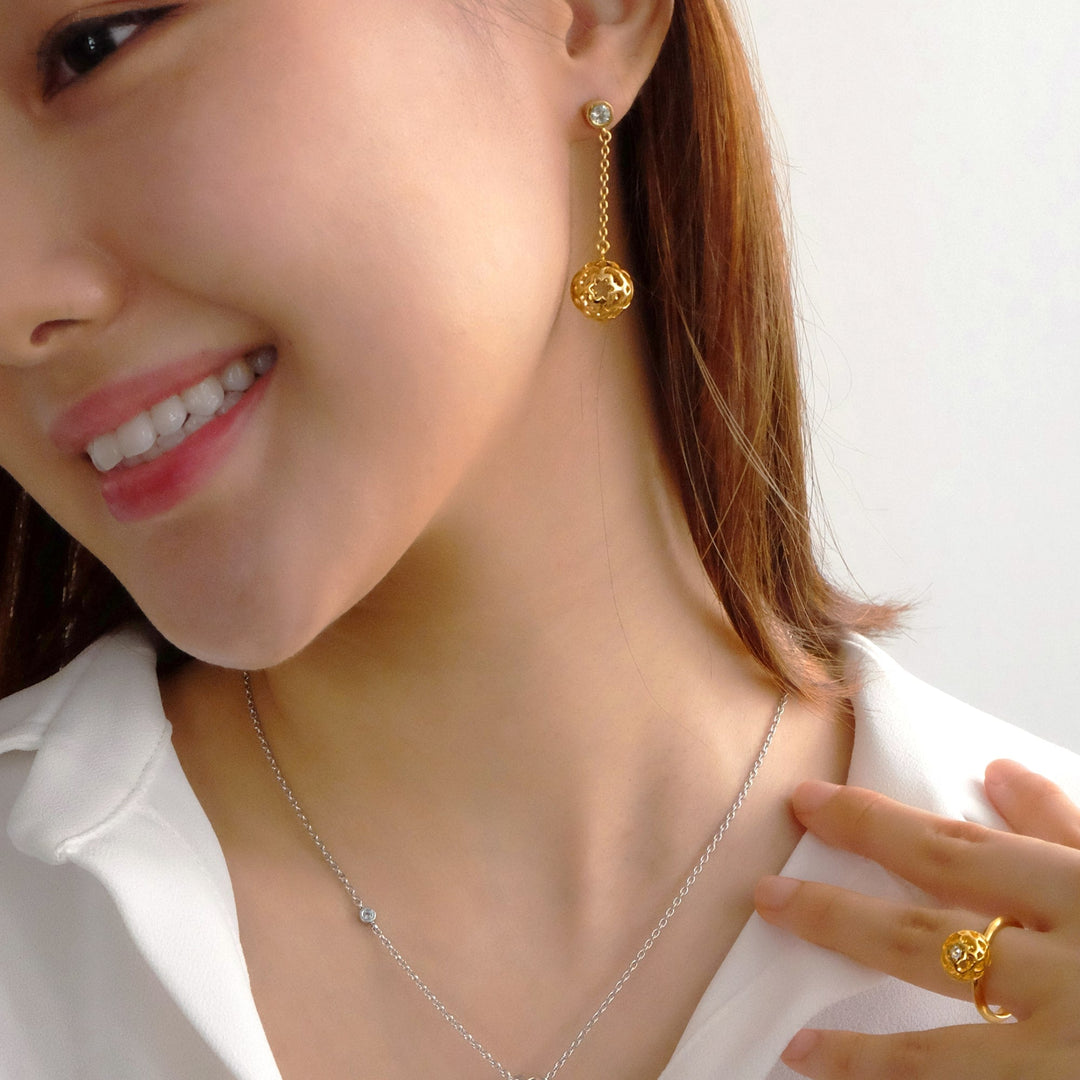Risis Peranakan Spheres Dangling Earrings with White Topaz (G) - A smiling person wears a gold dangle earring with a filigree sphere, a delicate necklace with a small gemstone, and a matching intricate gold ring on their finger.