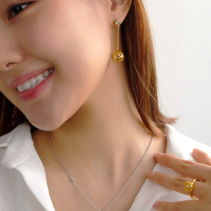 Risis Peranakan Spheres Dangling Earrings with White Topaz (G) - A smiling person wears a gold dangle earring with a filigree sphere, a delicate necklace with a small gemstone, and a matching intricate gold ring on their finger.