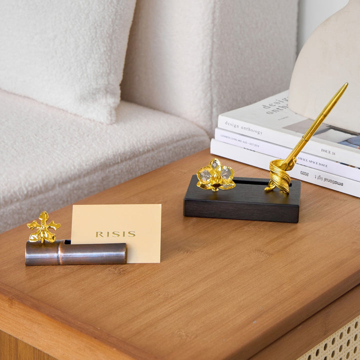 Risis Phalaenopsis Orchid Pen And Name Card Holder - Elegant gold-colored pen with a spiral design rests on a stand, beside a clear, crystal-like flower ornament and a small gold flower beside a metallic cylinder.