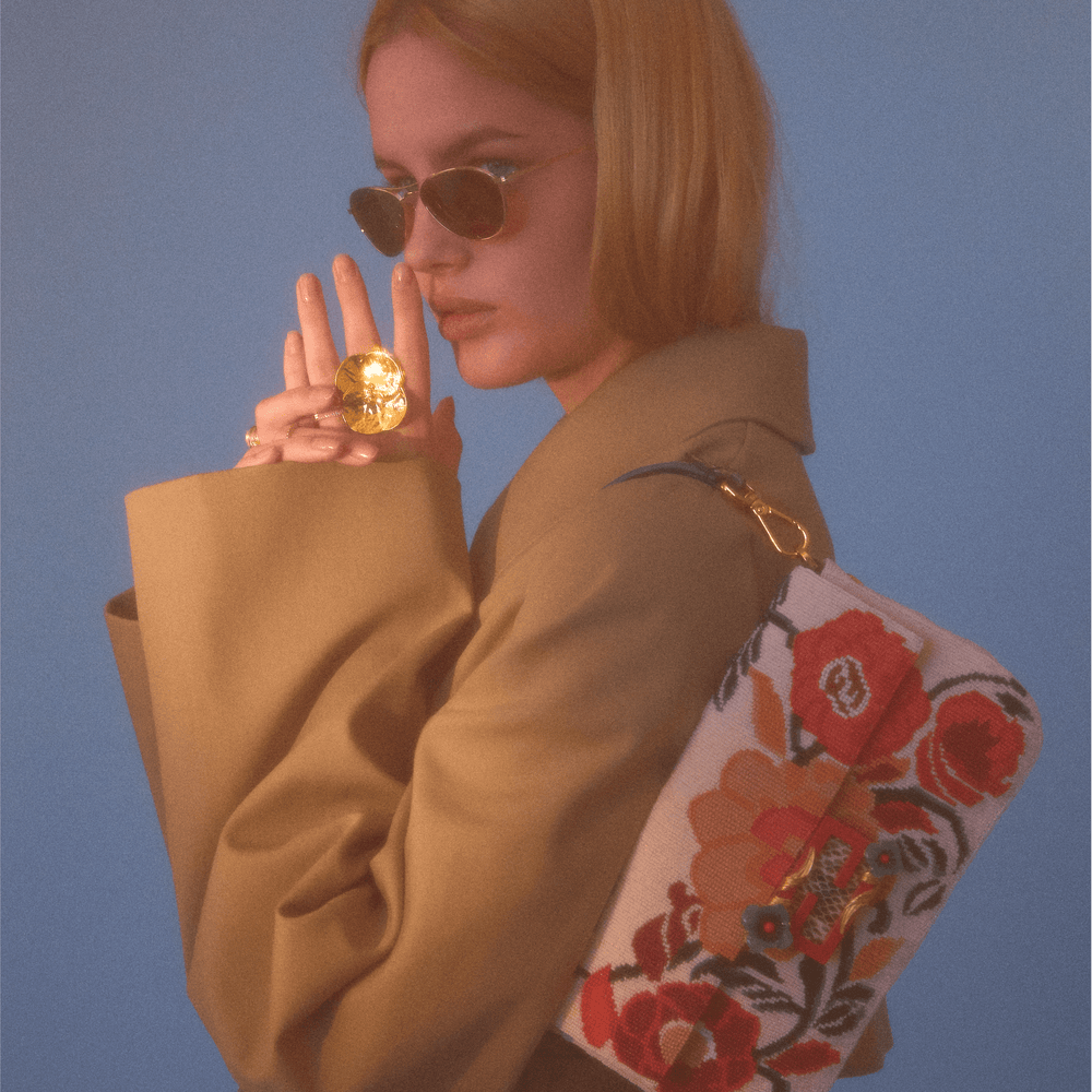 Risis Promesse Ring (G) - A person showcases a large, shiny golden ring on their finger, complemented by a colorful floral patterned purse with a noticeable texture on their shoulder.