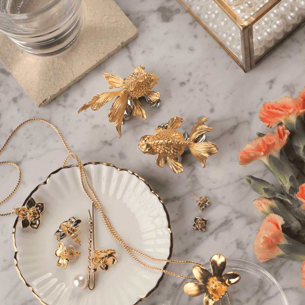 Risis Ranchu Goldfish Name Card Holder (Small) - Golden jewelry with intricate fish and floral designs is artfully displayed on a white scalloped plate, evoking elegance and sophistication.