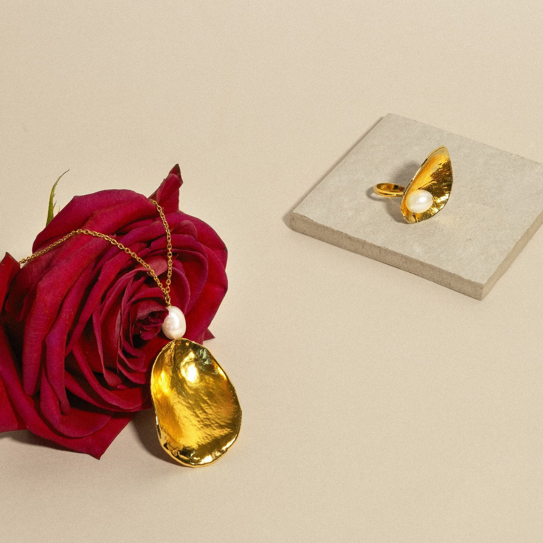 Risis Rose Petal Ring with Pearl - A gold pendant with a textured surface and a pearl accent dangles from a chain, entwined with a lush, deep red rose, next to a pearl-embellished gold ring on a stone tile.