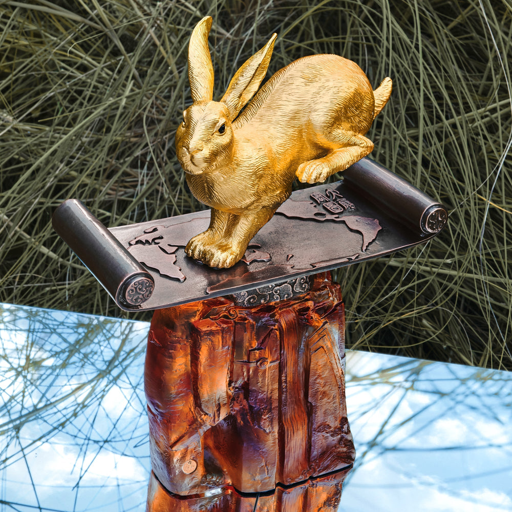 Risis The Magnificent Celestial - A golden rabbit sculpture stands atop an ornately carved dark metal cylinder resting on a polished amber-hued base.
