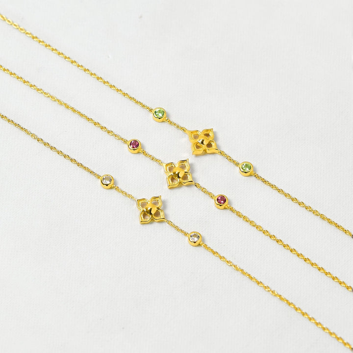 Risis Timeless Peranakan Bracelet With Peridot - A golden necklace with a bright yellow pendant and matching earrings are arranged on and around two stone blocks, complemented by delicate pink flower blossoms.