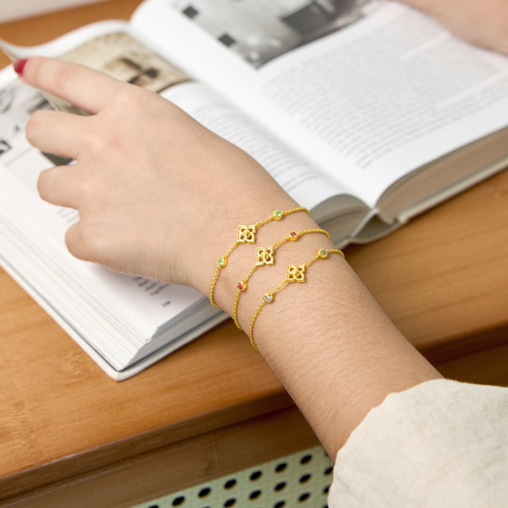 Risis Timeless Peranakan Bracelet With Pink Tourmaline - A golden bracelet with intricate honeycomb-shaped links adorned with tiny colored gems wraps delicately around a person's wrist resting on an open magazine.
