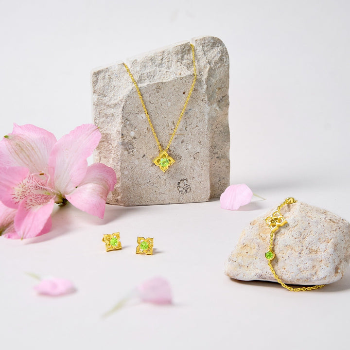 Risis Timeless Peranakan Earrings With Peridot - Gold jewelry with green gemstones is displayed amidst pink flowers and stones. Earrings, a necklace, and bracelet showcase vibrant colors and exquisite details.