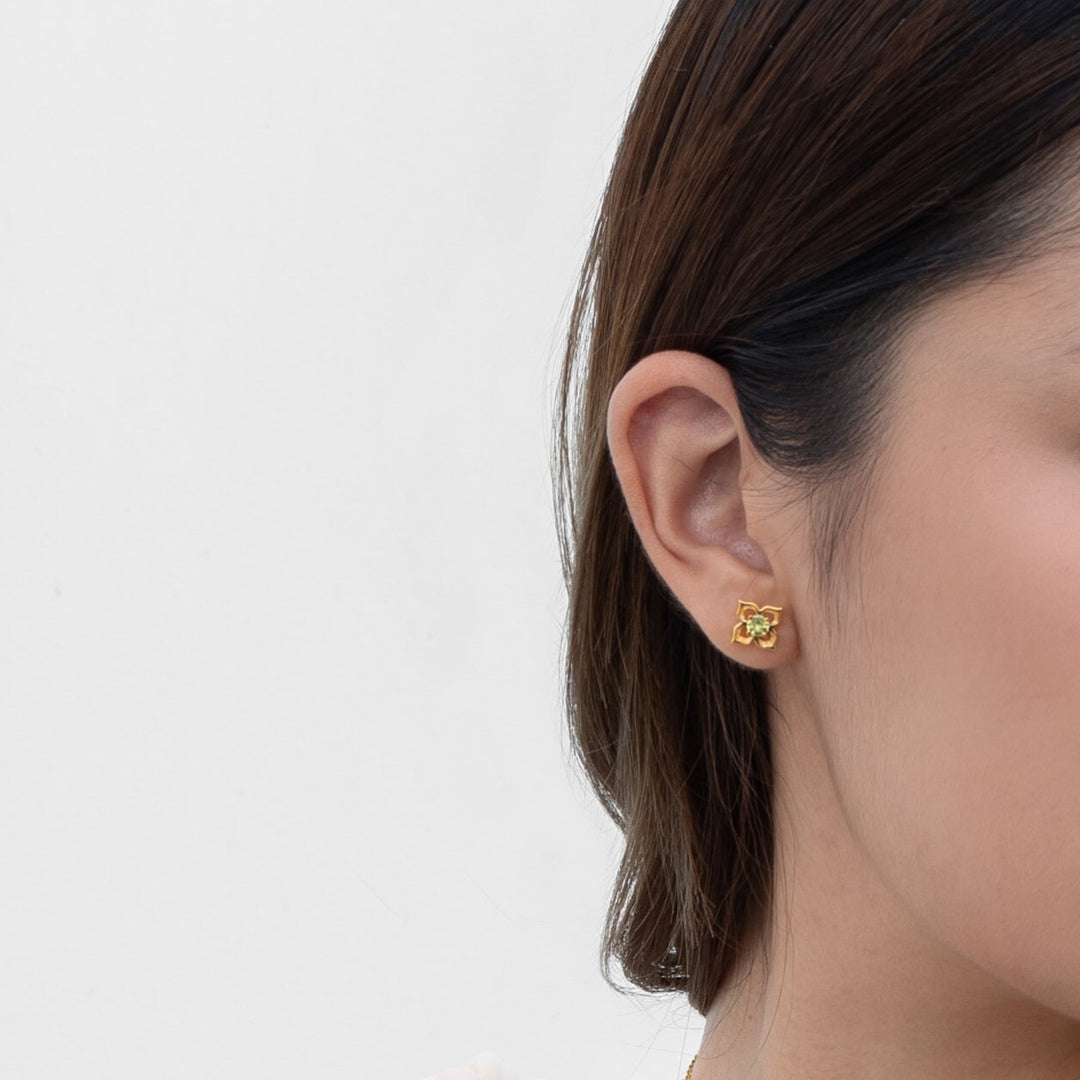 Risis Timeless Peranakan Earrings With Peridot - A gold earring with a flower design and a central green gemstone is worn on a person's ear, displaying an elegant, simplistic style.