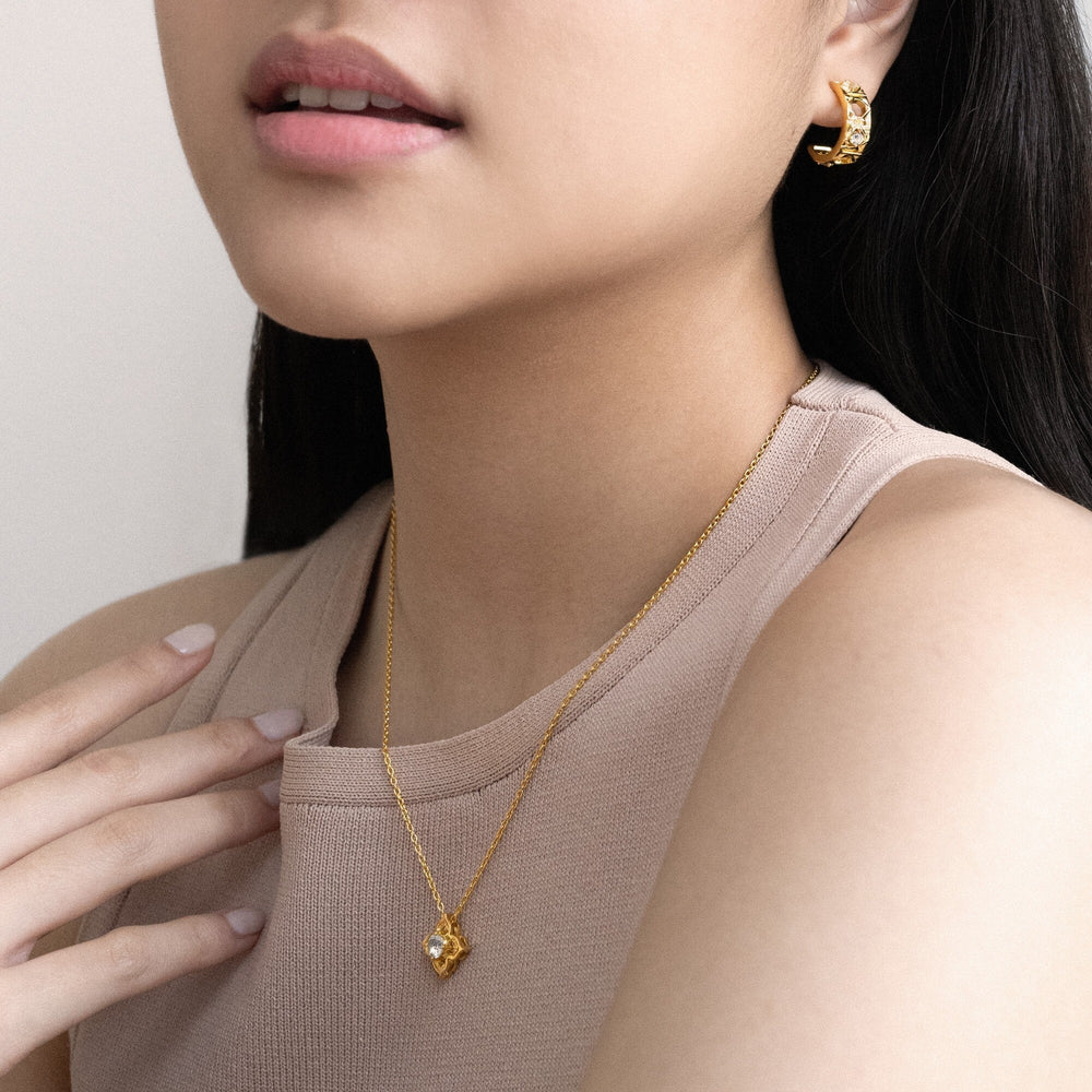 Risis Timeless Peranakan Small Necklace with White Topaz - Golden hoop earrings and matching pendant necklace adorn a person. The earrings have a twisted texture, while the necklace features a clear, faceted gemstone inside a gold setting.