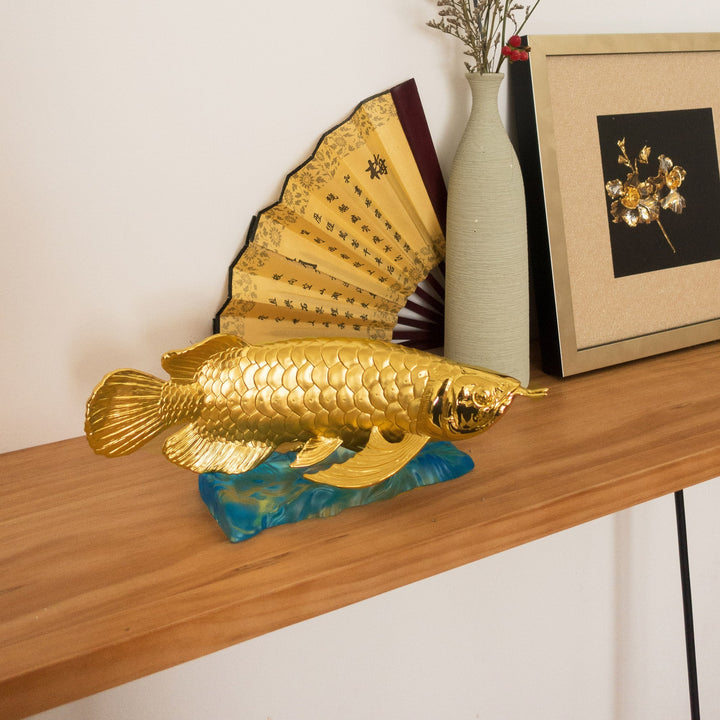 Risis Treasure Arowana - A golden fish sculpture with detailed scales rests on a blue wave-like base, exuding an elegant and ornate aesthetic.