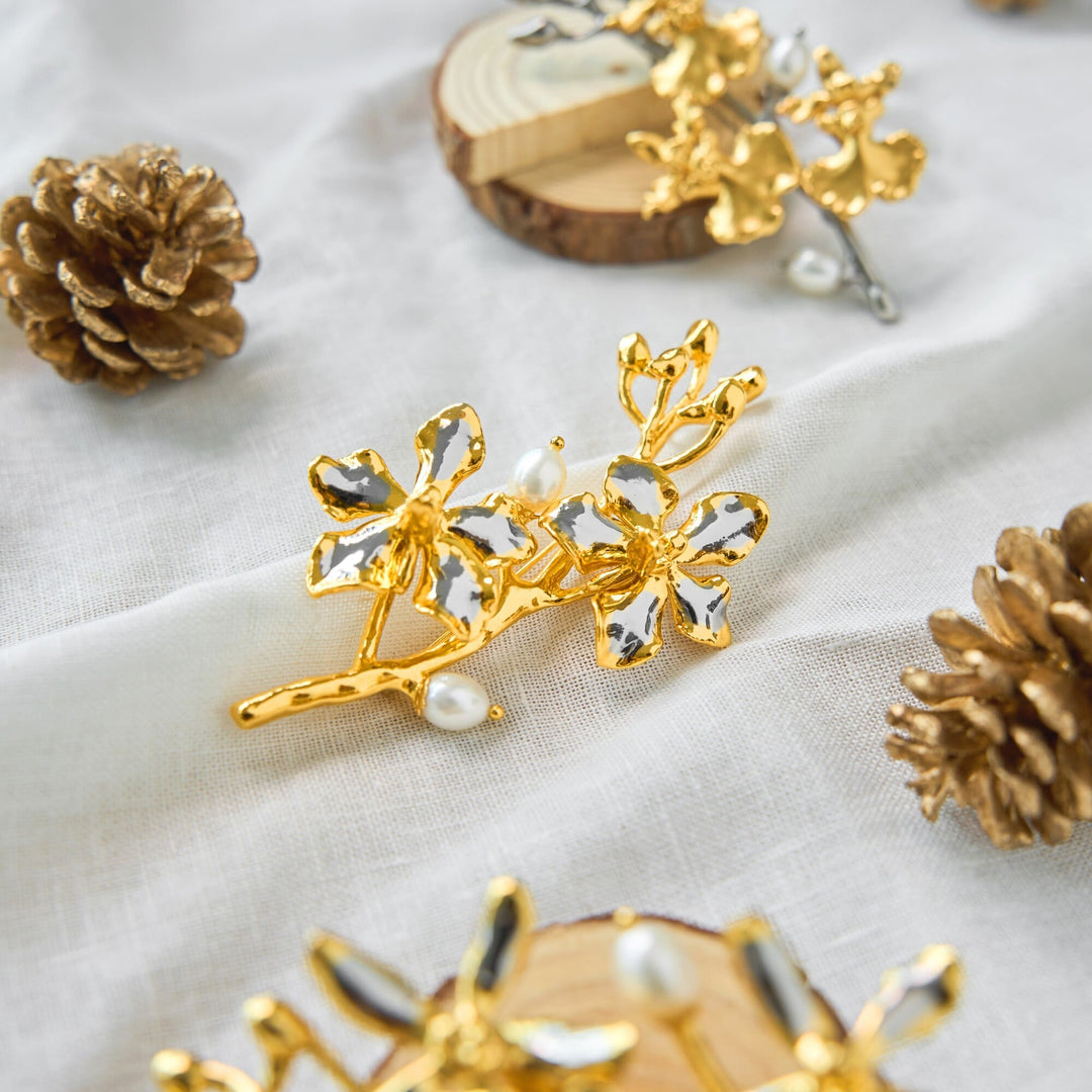 Risis Vanda Symphony Brooch with Pearls - A golden hair accessory adorned with glossy, flower-shaped elements and delicate pearl embellishments rests elegantly against a soft, textured surface.