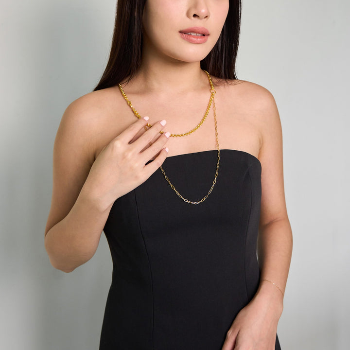 Risis Vanda Symphony Necklace with Pearls - A person is adorned with a gold layered necklace featuring varied chain designs. Their hand, with matching rings, gently touches the necklace, accentuating its elegance against a black dress.