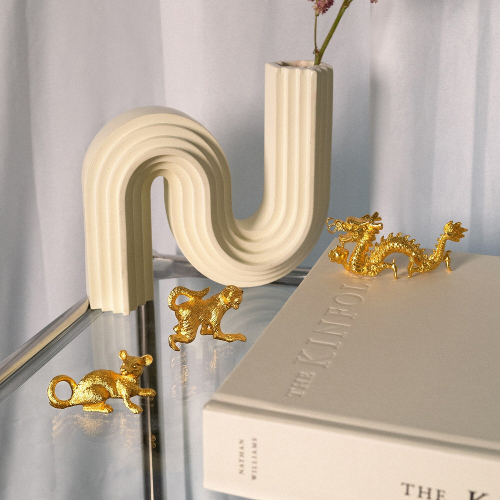 Risis Zodiac Monkey Figurine - Three golden animal figurines—a kangaroo, a monkey, and a dragon—are arranged on a reflective surface next to an arched white object and a book.