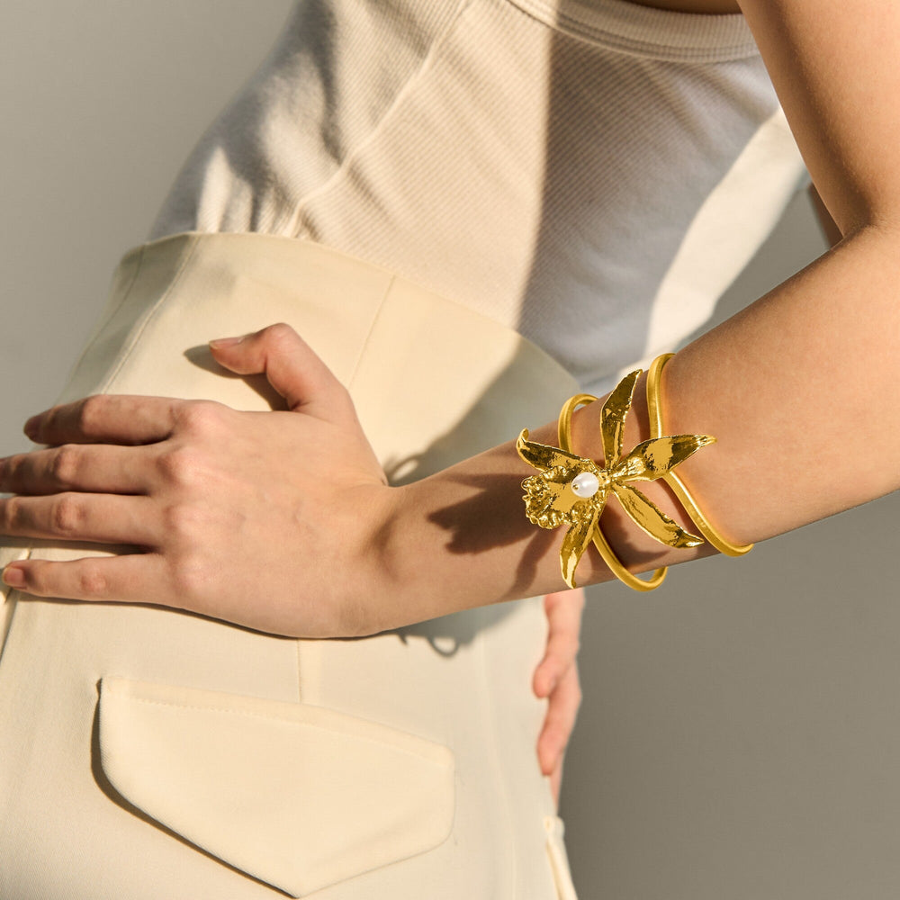 Risis 1974 Brassidium Cuff Bangle - A gold bracelet with a floral motif and a central pearl adorns a person's wrist, complementing a cream outfit.