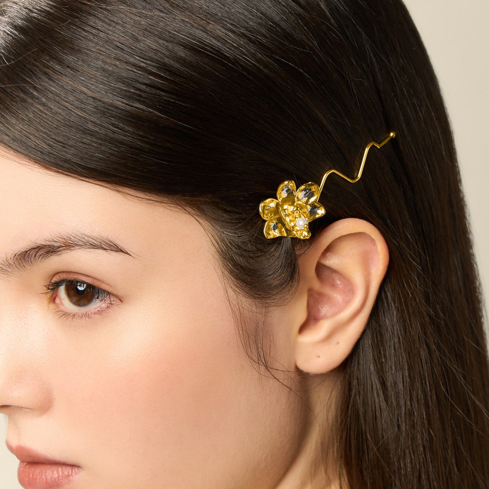 Risis 1974 Doritaenopsis Hair Clip - A golden floral hairpin with glittering petals adorns the hair, its wavy stem elegantly tucked behind a woman's ear, imparting a touch of luxury.