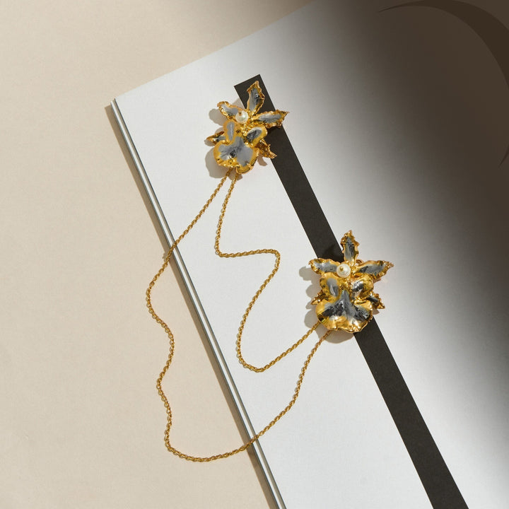 Risis 1974 Oncidium Chain Brooch - Two ornate, gold-tone brooches with flower designs, accented with white enamel and pearl-like details, connected by a delicate chain, displayed on paper with contrasting lines.