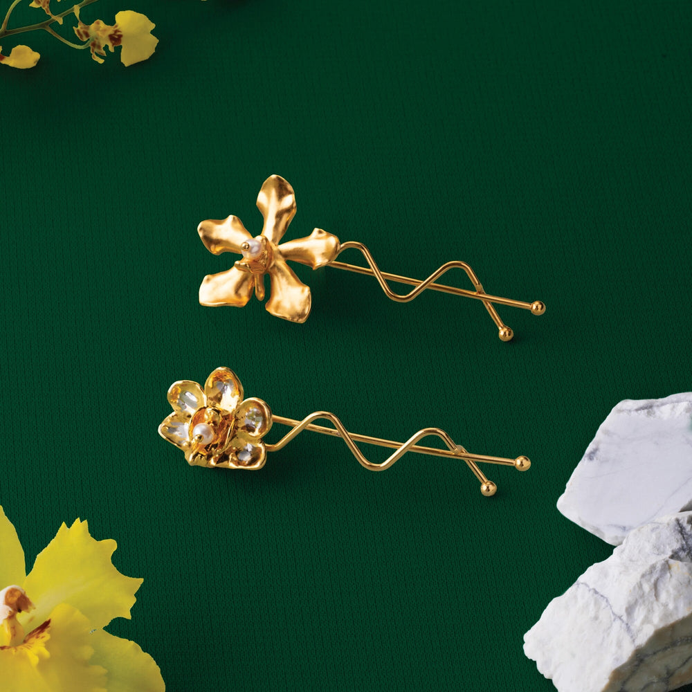 Risis 1974 Vanda Hair Clip - Two golden earrings, shaped like abstract flowers with a lustrous finish, are displayed on a dark green surface.