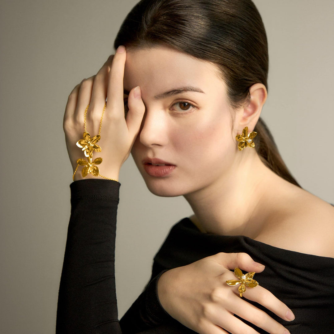 Risis Aeridovanda Blooms Ring - A woman showcases golden jewelry with floral motifs; she wears an ornate bracelet, earrings, and ring with amber-like gem petals, all exuding elegance and luxury.