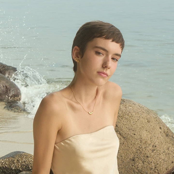 Risis Ascocenda Garden Necklace - A person with a pixie haircut wears gold earrings and a delicate necklace. The jewelry exudes simplicity and elegance against the individual's neutral attire.