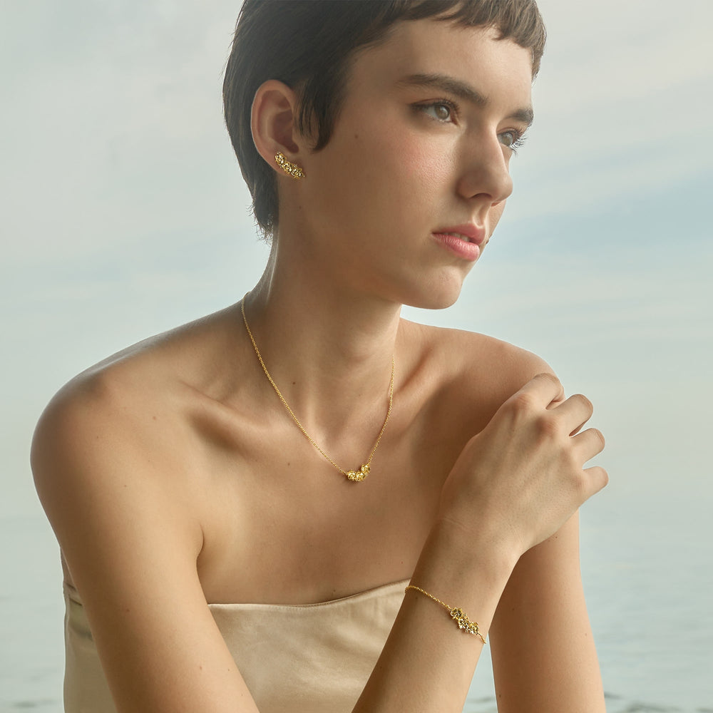 Risis Ascocenda Garden Necklace - A woman dons delicate gold jewelry, including textured earrings, a matching pendant necklace, and a bracelet, all showcasing elegance and simplicity.