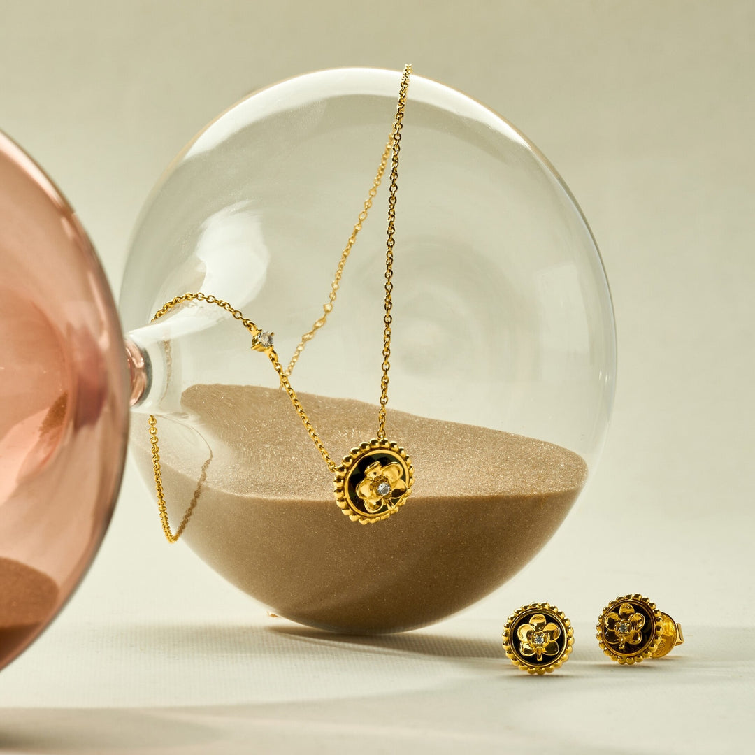 Risis Beaded Ascocenda Earrings - A golden pendant with a central gemstone hangs inside a transparent orb partially filled with sand, accompanied by two matching golden stud earrings nearby.