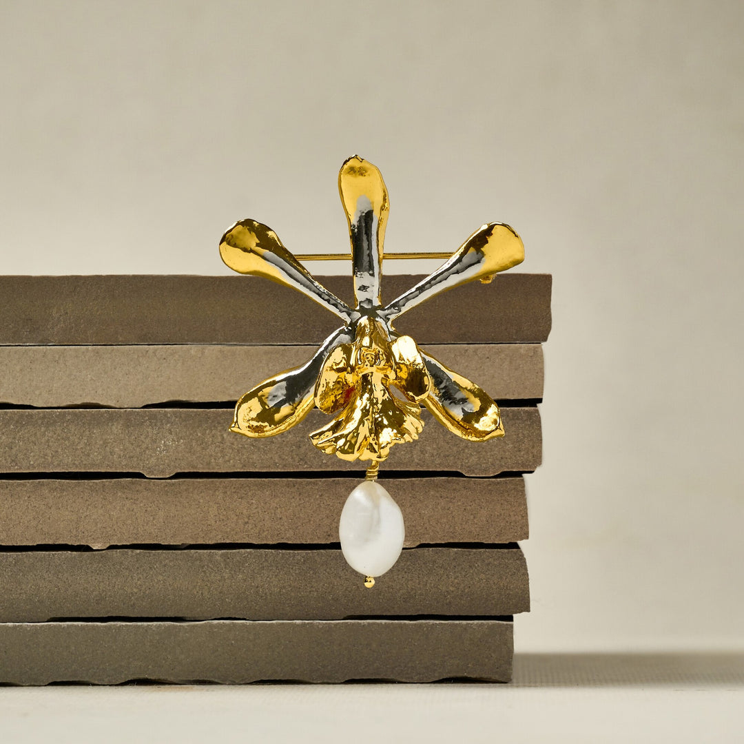 Risis Encyclia Tampensis Orchid Pearl Brooch - A shiny gold-toned orchid brooch with reflective surfaces and a dangling white pearl is positioned on dark horizontal layers.