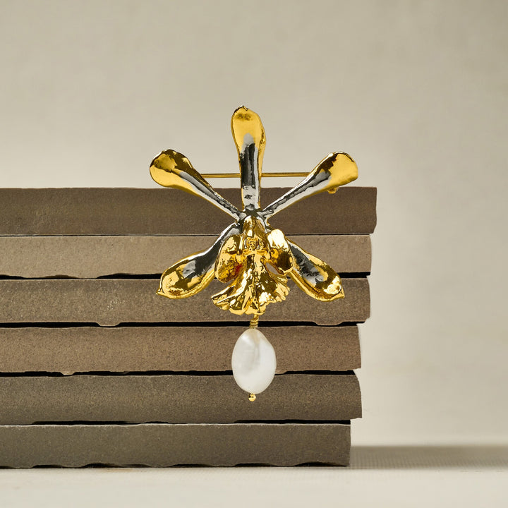 Risis Encyclia Tampensis Orchid Pearl Brooch - A shiny gold-toned orchid brooch with reflective surfaces and a dangling white pearl is positioned on dark horizontal layers.