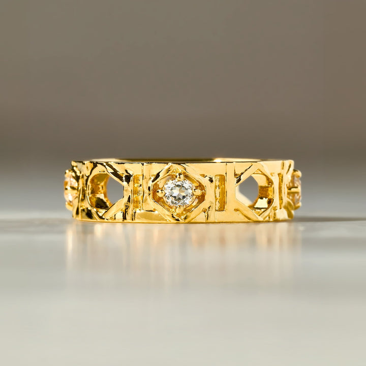 Risis Entwined Rattan Ring - A golden ring with intricate cut-out patterns and a central diamond rests on a reflective surface, showcasing its ornate design and shimmering jewel.