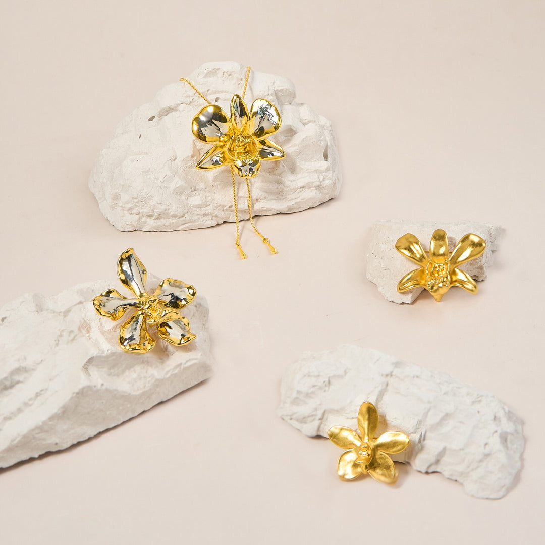 Risis Aeridovanda Vieng Ping Orchid Brooch/Pendant (G) - Four golden flower-shaped jewelry pieces rest on jagged white surfaces, reflecting light with a metallic sheen, giving off an elegant aura.