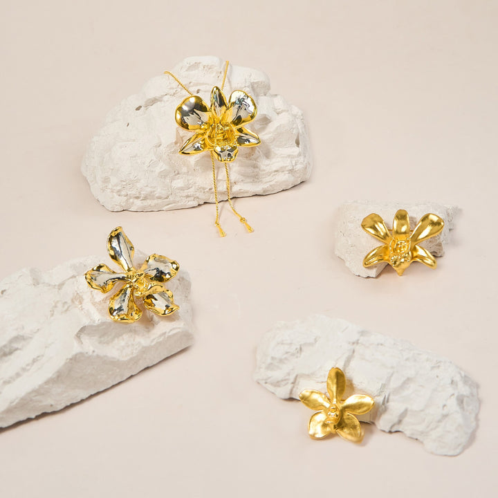 Risis Aeridovanda Vieng Ping Orchid Brooch/Pendant (G) - Four golden flower-shaped jewelry pieces rest on jagged white surfaces, reflecting light with a metallic sheen, giving off an elegant aura.