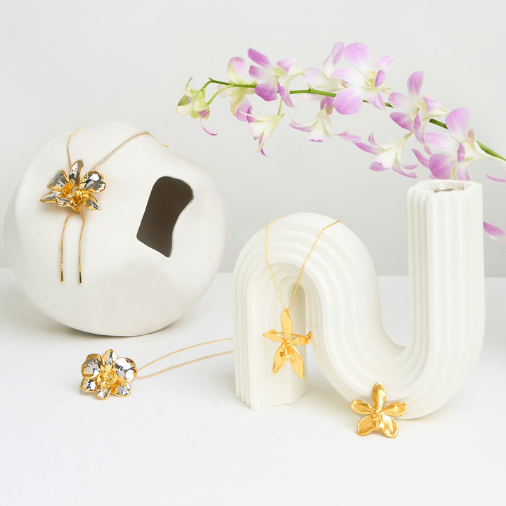 Risis Phalaenopsis Cornu Cervi Orchid Necklace (G) - Golden flower-shaped jewelry pieces with elongated petals are displayed atop and beside white, sculptural stands, accented by delicate purple orchids draped in the background.