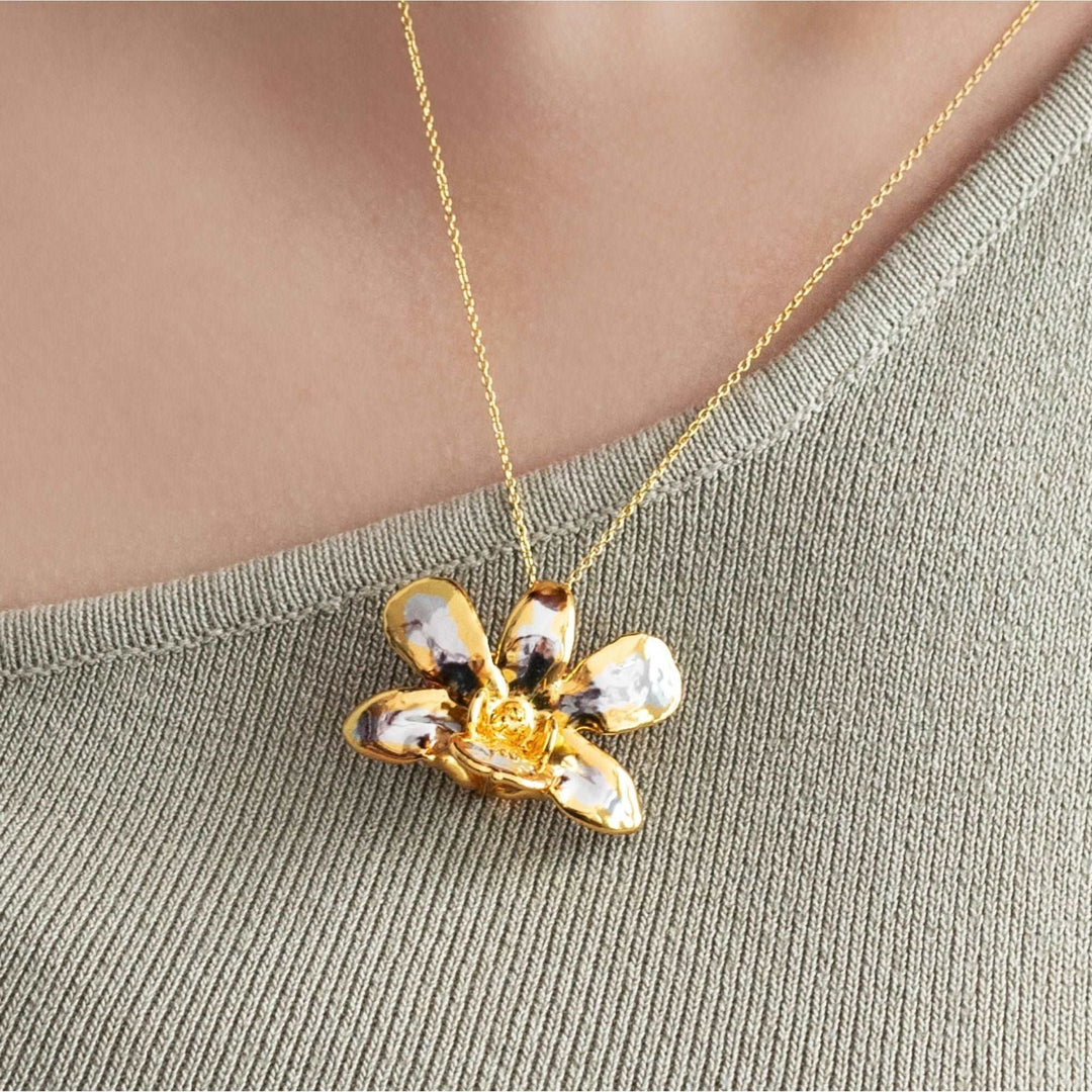 Risis Dendrobium Thong Chai Orchid Necklace (PG) - A golden necklace with a shiny, reflective flower pendant, featuring a lustrous finish and intricate petal details, worn against a fabric background.