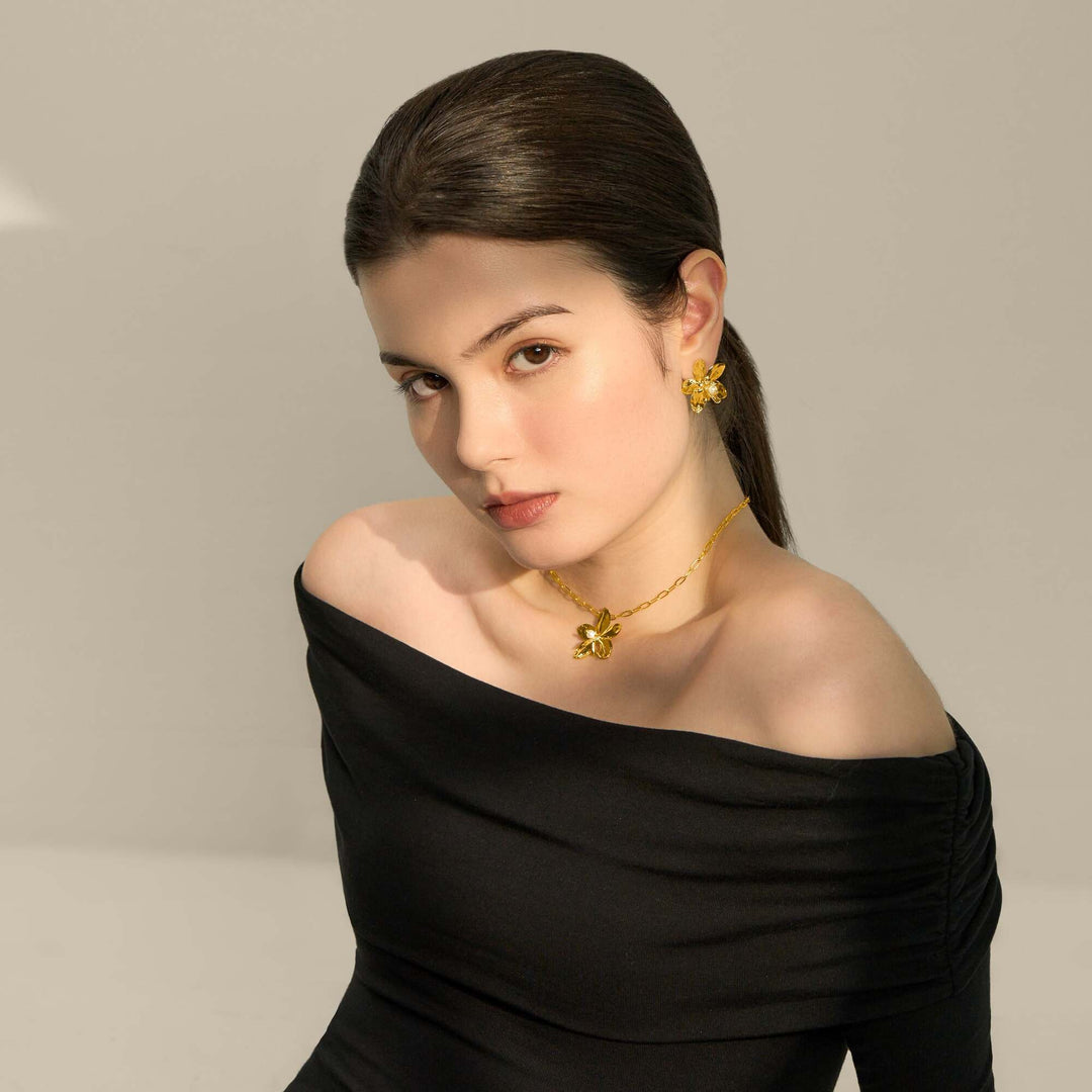 Risis Aeridovanda Blooms Earrings - A woman is wearing golden floral jewelry; the necklace and earrings feature intricate petals and an accent gem, contrasting with her elegant black attire.