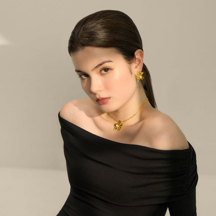 Risis Aeridovanda Blooms Earrings - A woman is wearing golden floral jewelry; the necklace and earrings feature intricate petals and an accent gem, contrasting with her elegant black attire.