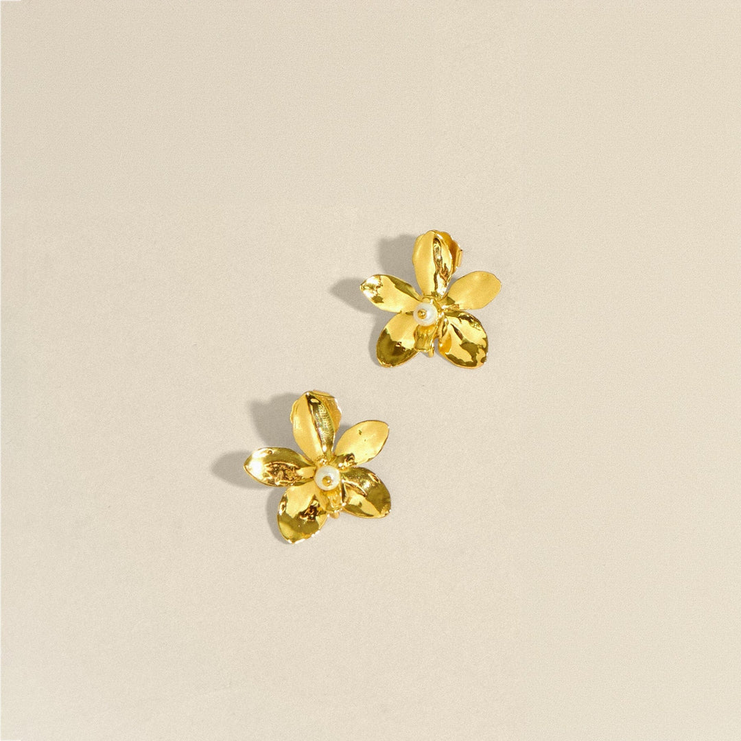 Risis Aeridovanda Blooms Earrings - A pair of golden earrings shaped like five-petaled flowers with a pearl at the center, lying on a neutral surface.