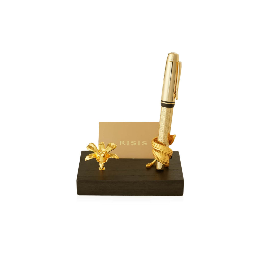 Risis Dendrobium Li Min Eng Orchid Pen And Name Card Holder - A golden pen with textured detailing stands vertically, intertwined with a sculptural golden ribbon, beside a delicate gold orchid, all presented on a sleek, dark wooden base.