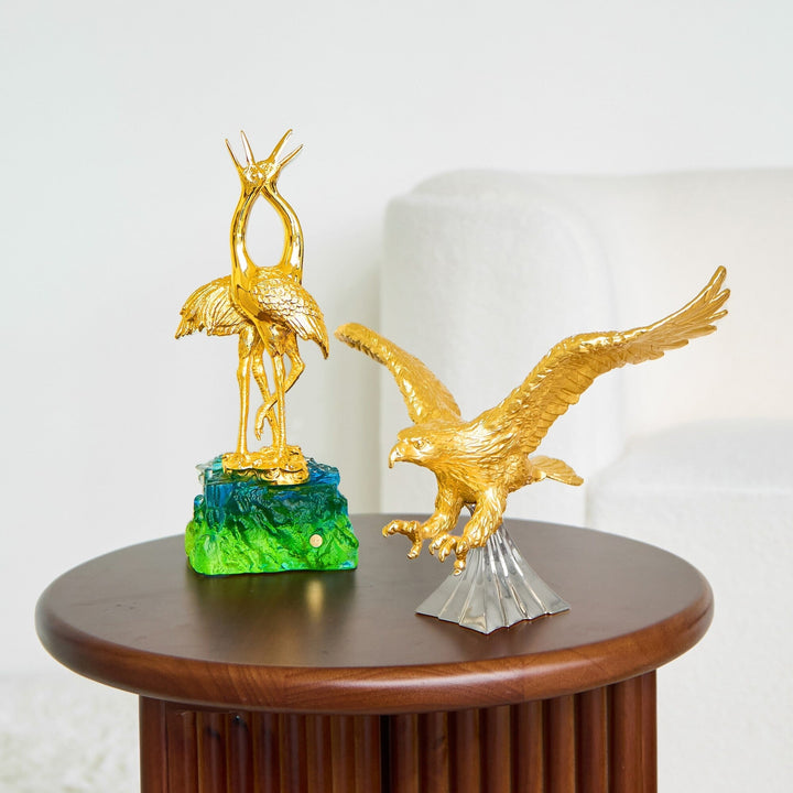 Risis Double Crane on Liu Li - Two golden bird sculptures; one stands majestically atop a vibrant green base, while the other appears to be taking flight from a silver pedestal.