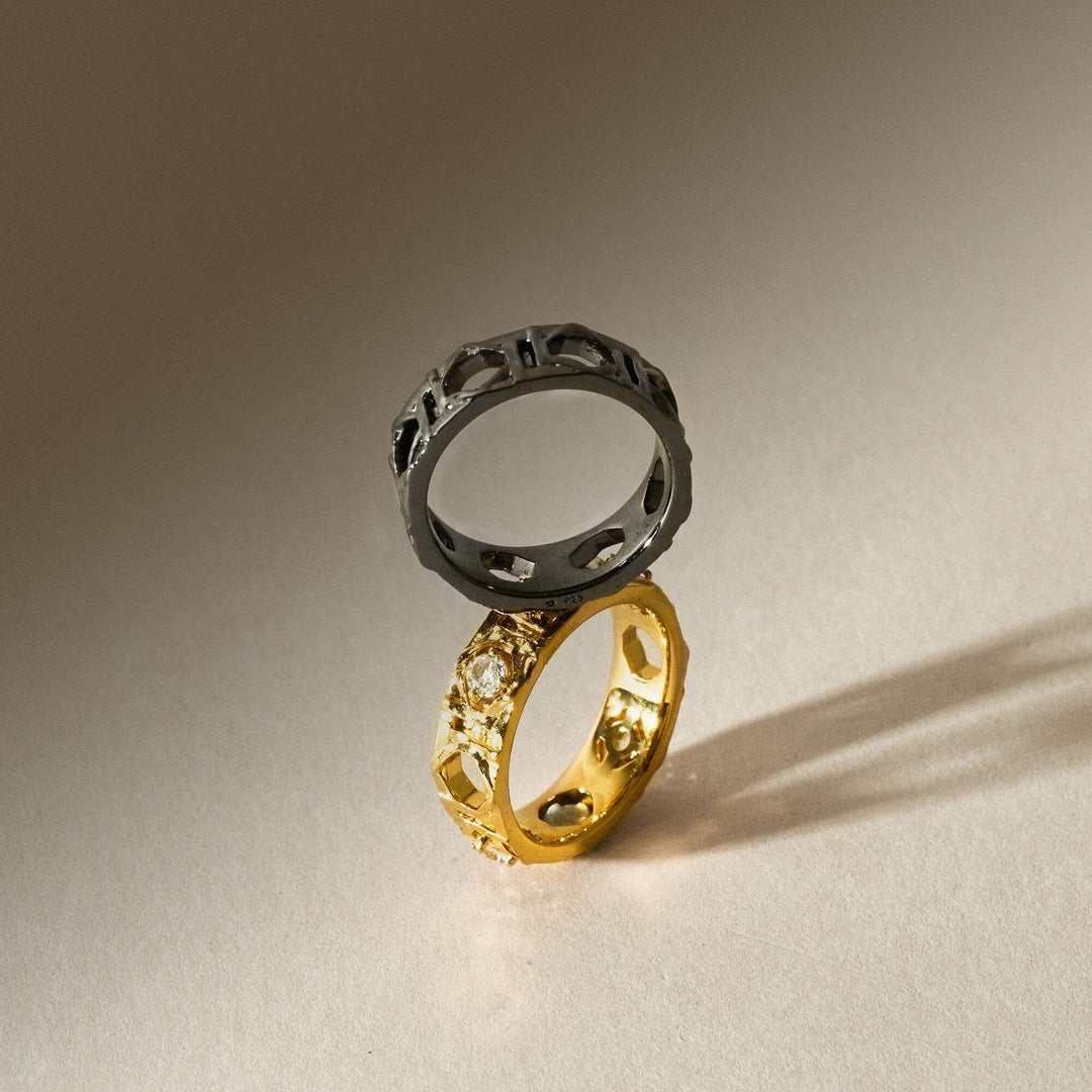 Risis Entwined Rattan Ring Brh - Two rings, one silver with geometric cutouts, the other gold with embedded stones, intersect delicately on a smooth surface, their shadows elongating behind them.