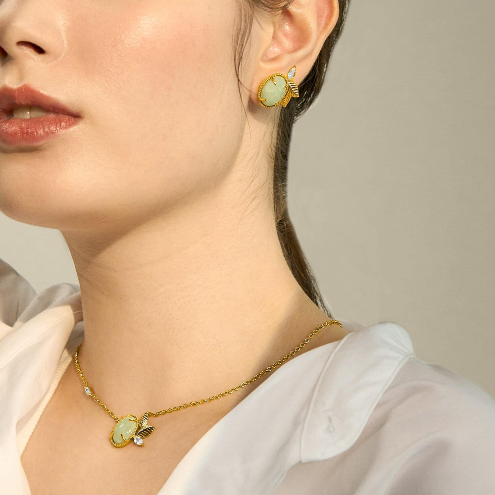 Risis Jadeite Prairie Necklace - A woman displays gold-colored jewelry: round, olive gemstone earrings with leaf motifs and a matching pendant necklace featuring a bumblebee adorned with small, glittering accents.