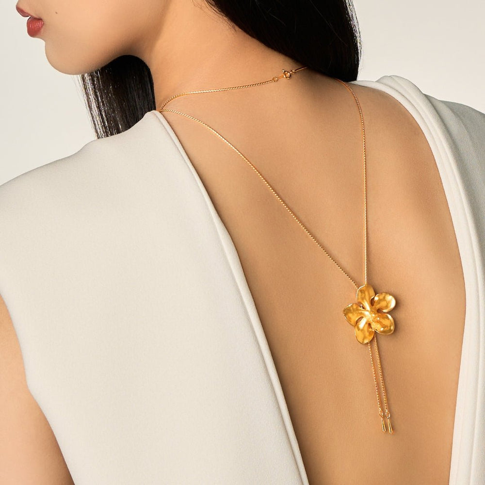 Risis Kagawara Firebird Orchid Slider Necklace (G) - A gold pendant resembling a stylized, four-petaled flower dangles on a thin chain, with two delicate tassels hanging from its bottom.
