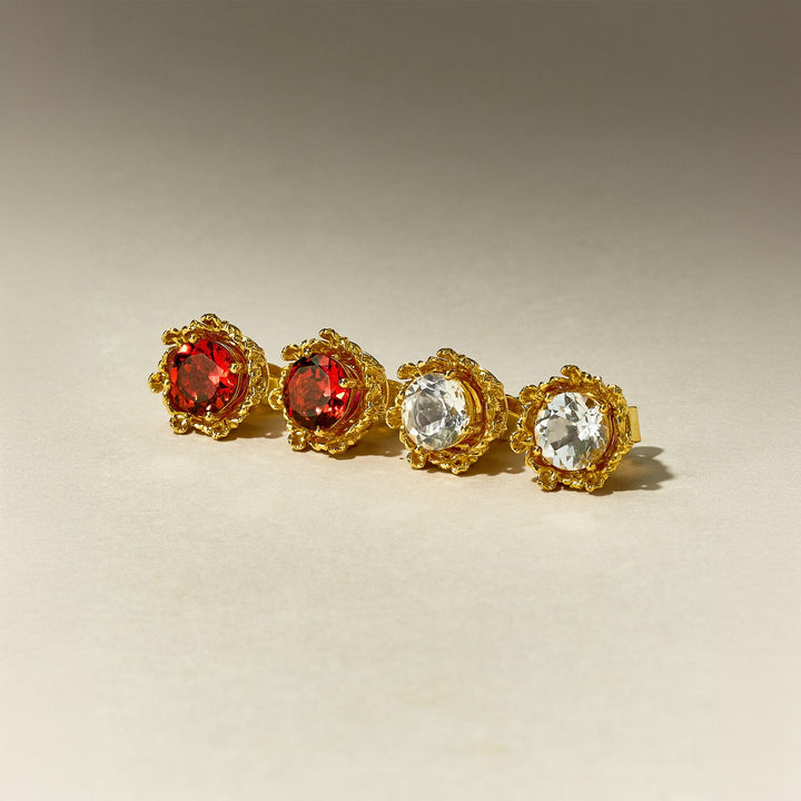 Risis Majestic Vanda Earrings With Red Garnet - Four ornate rings with gold filigree settings are arrayed in a row; two feature deep red gemstones while the other two showcase clear, sparkling gems.