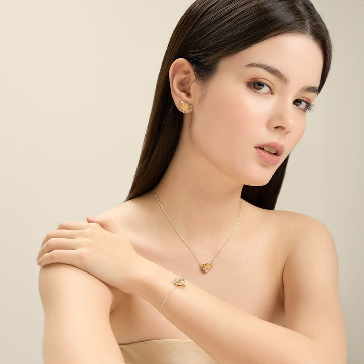 Risis Magnolia Blossom Necklace - A woman adorned with gold jewelry with gemstone accents—stud earrings, a pendant necklace, and a bracelet—reflects elegance and simplicity.