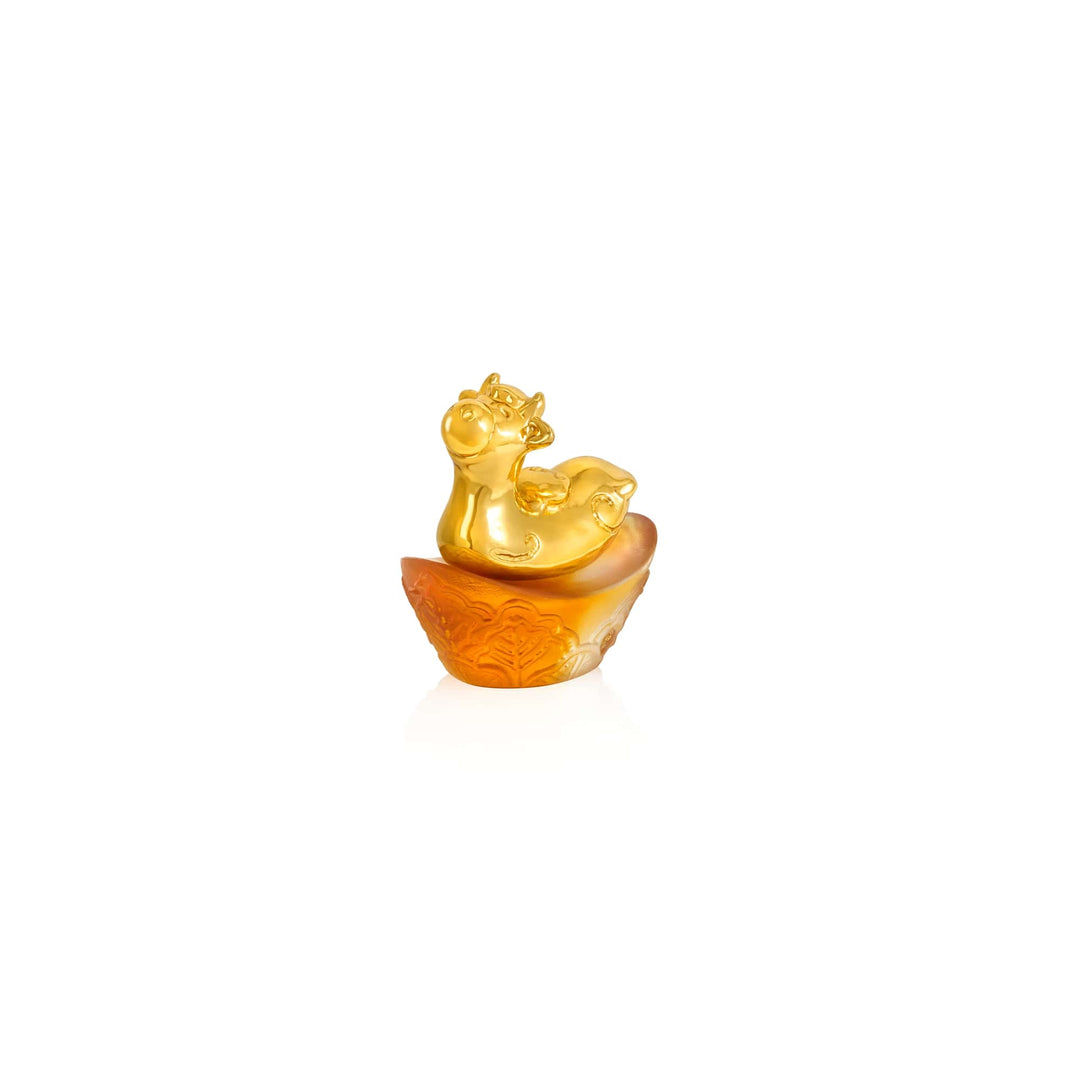 Risis Mini Celestial – Ox on Amber Liu Li - A golden figurine of a cheerful, cartoon-like cow playfully reclined atop an orange, intricately carved base, possibly symbolizing prosperity or good fortune.
