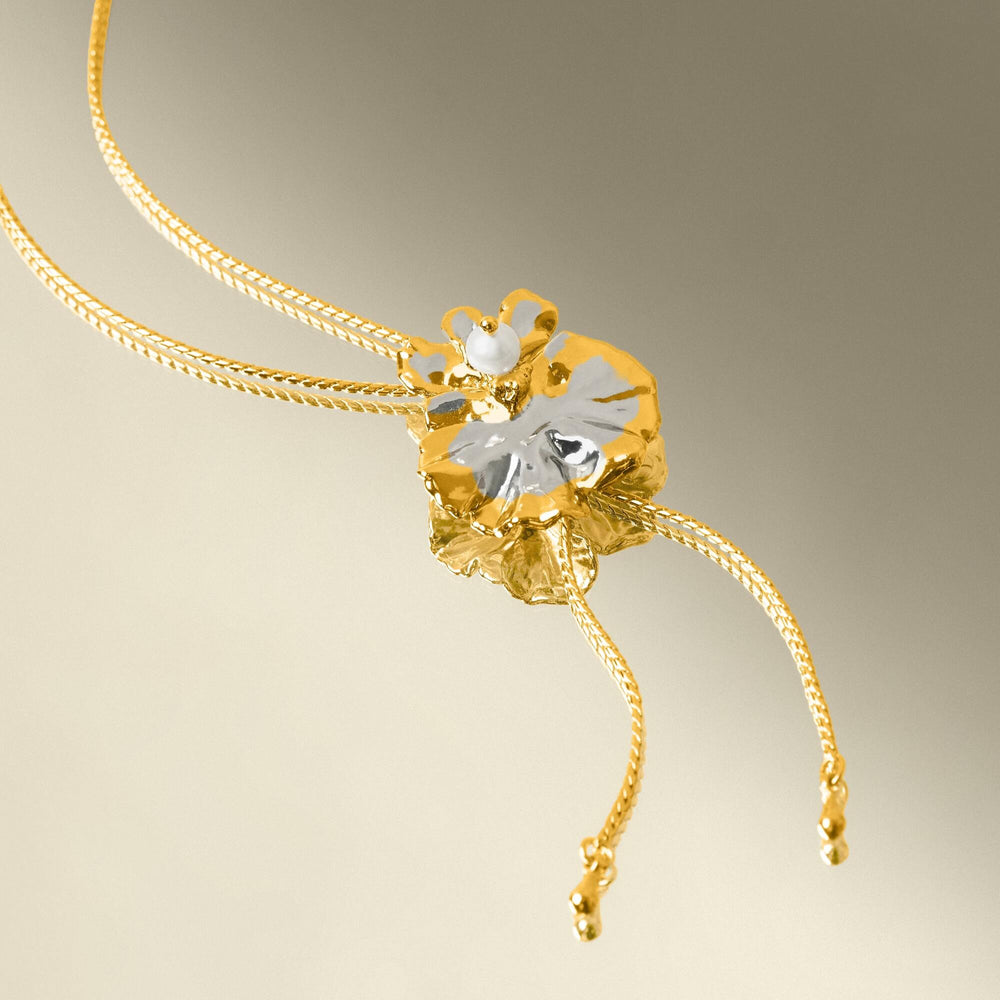 Risis Oncidium Coco Drop Pearl Slider Necklace Pg - A golden necklace with a flower-shaped pendant, featuring a central pearl and delicate petals, accented with a silver leaf, on thin intertwined chains.